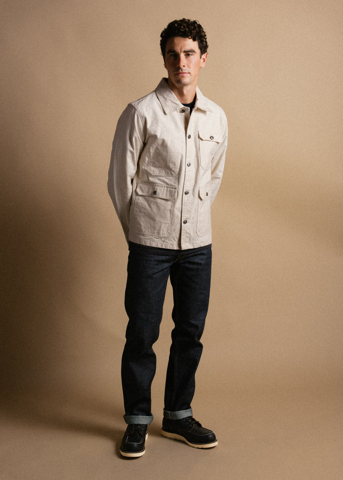 Man in beige jacket and dark jeans stands against a neutral background, hands behind back, exuding casual style and confidence.