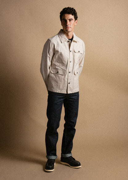 Man in beige jacket and dark jeans stands against a neutral background, hands behind back, exuding casual style and confidence.