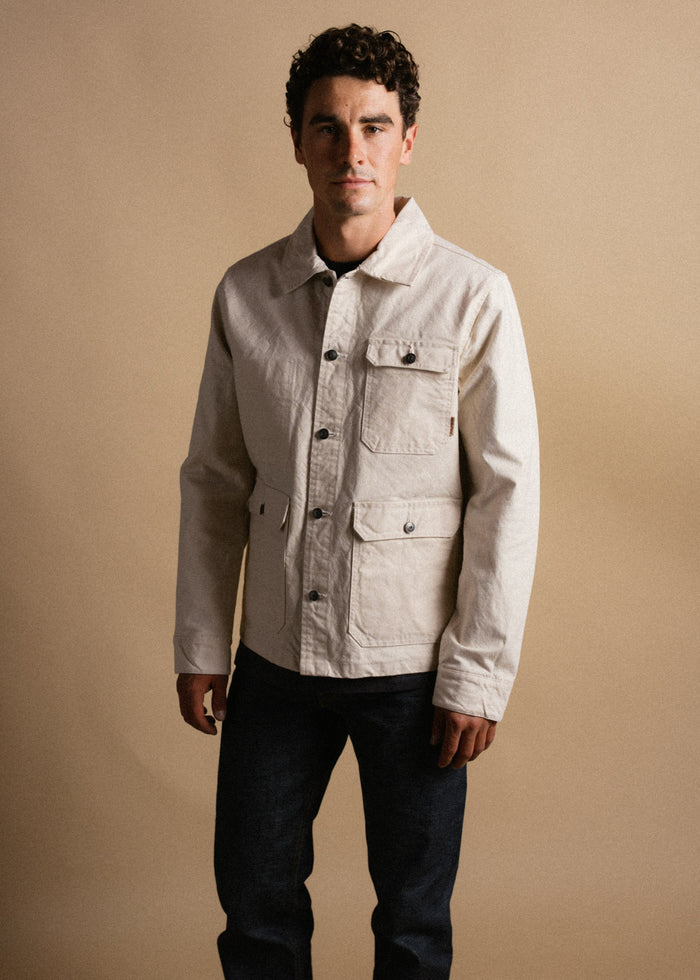 Man in beige jacket and dark jeans stands against a plain background, looking forward with a neutral expression.