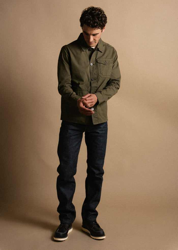 Man in olive jacket and dark jeans stands against beige background, adjusting cuffs. Casual, stylish look with focus on outfit.