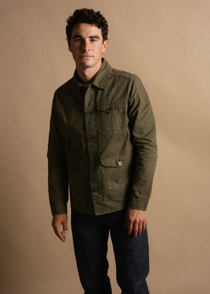 Man in olive jacket and jeans stands against a beige background, exuding a casual yet stylish look with a confident expression.