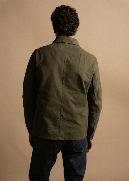 Man with curly hair in olive jacket and jeans stands facing away against a beige background, highlighting casual fashion style.