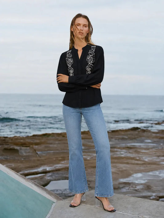 A woman stands on a rocky beach wearing a black long-sleeve blouse with ornate white embroidery down the front placket and light blue flare jeans.