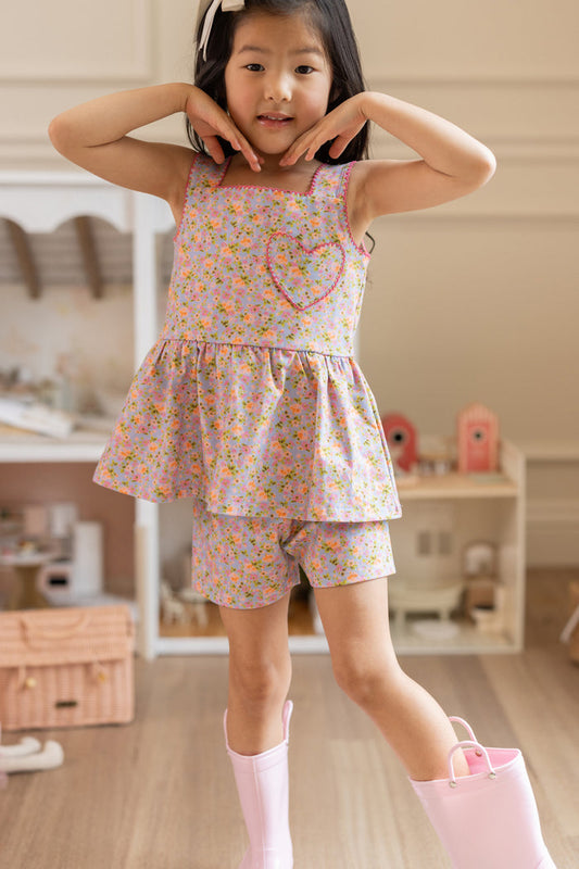 A young girl smiles indoors, posing in the Ivy City Co Mini Blair Play Set in Vibrant English Garden with pink rain boots. She wears a white bow in her hair and frames her face with her hands.