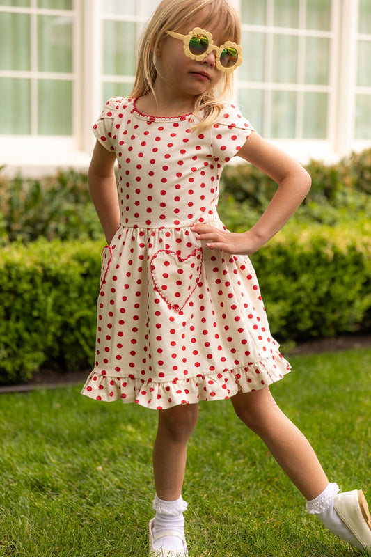 A young girl poses on grass in the Ivy City Co Mini Dream Play Dress in Red Polka Dot, featuring heart-shaped pockets and a ruffled hem. She wears yellow sunglasses, white socks, and shoes. Green hedges and windows complete the background.