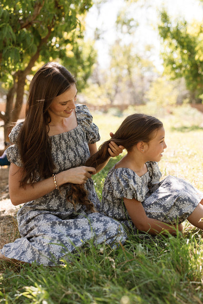 A woman sits on the grass braiding a young girl's hair, both smiling in a sunlit outdoor setting with trees behind them. They wear matching Ivy City Co Mini Lisa Dresses in Jolie Fleur, featuring puff sleeves and smocked bodices.