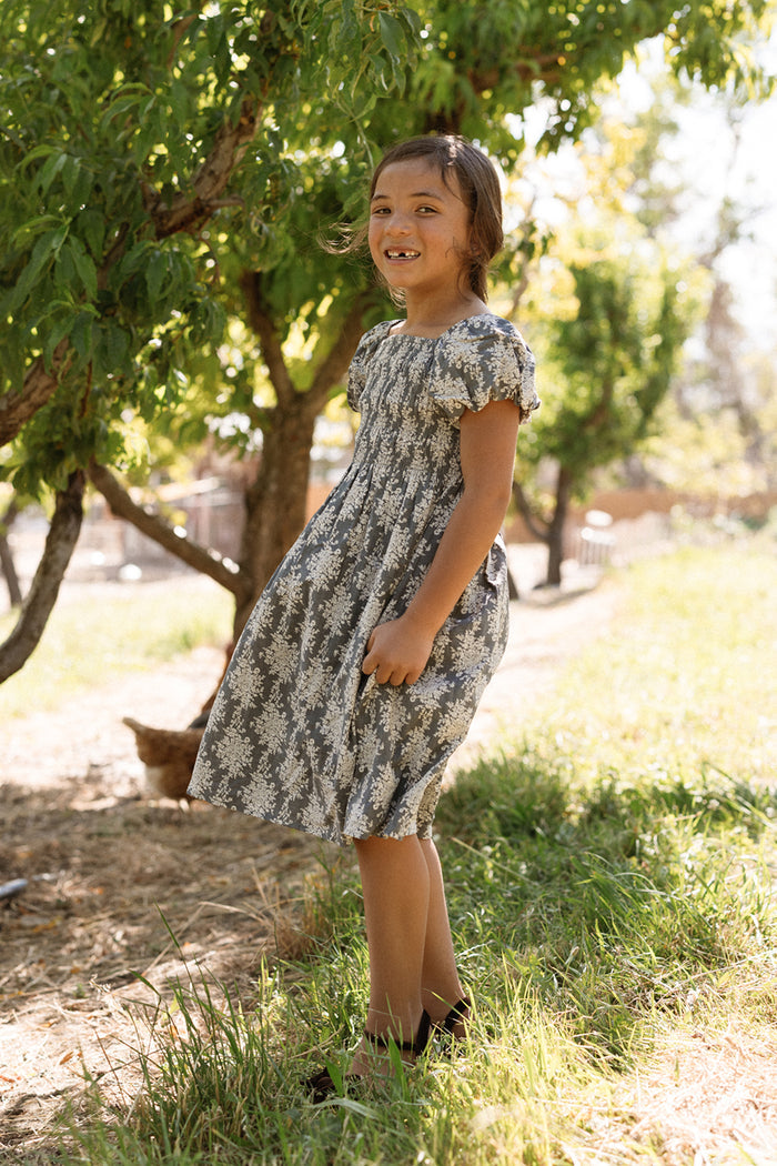 A young girl stands smiling outside on the grass near a tree on a sunny day, wearing the Ivy City Co Mini Lisa Dress in Jolie Fleur, which features a gray floral pattern, smocked bodice, and puff sleeves.