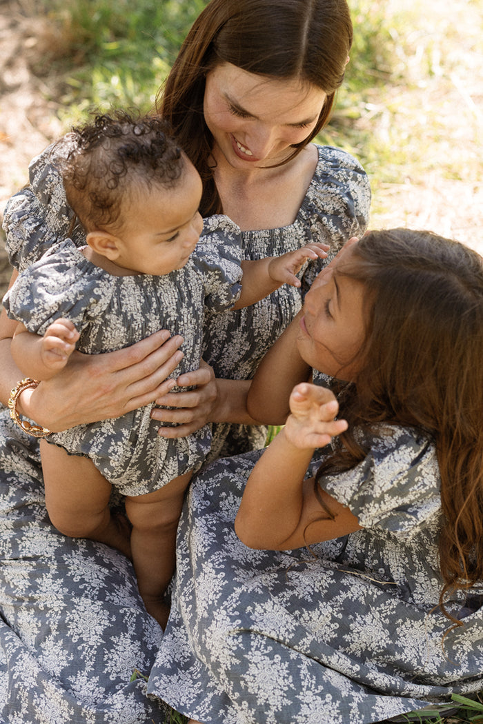 A woman, a baby, and a young girl sit together outdoors in the sunlight, smiling and interacting. All three wear coordinated Ivy City Co Mini Lisa Dresses in Jolie Fleur, featuring smocked bodices and charming puff sleeves adorned with a blue and white floral pattern.