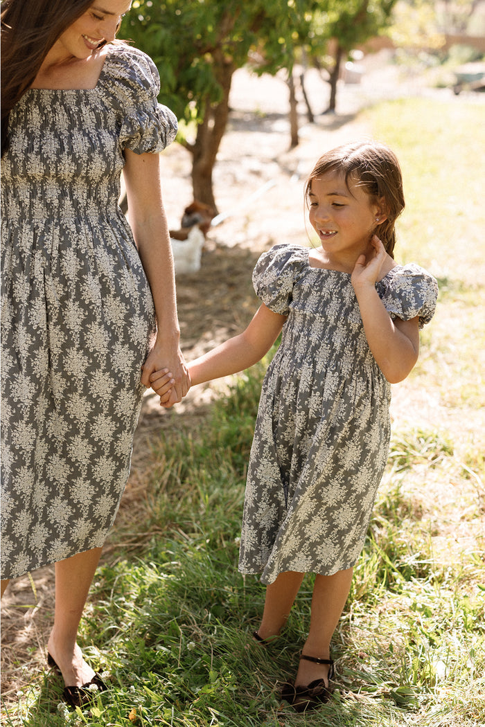 A woman and a young girl, both wearing matching Ivy City Co Mini Lisa Dresses in Jolie Fleur with puff sleeves and smocked bodices, hold hands and smile while walking outdoors on a sunny day. Trees and grass fill the background.