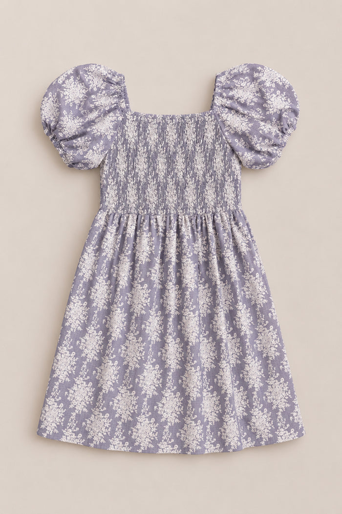 Introducing the Mini Lisa Dress in Jolie Fleur by Ivy City Co: a delightful lavender dress showcasing short puff sleeves and a smocked bodice, beautifully accented with delicate white floral patterns throughout. Displayed against a neutral background, the dress exudes whimsical elegance.