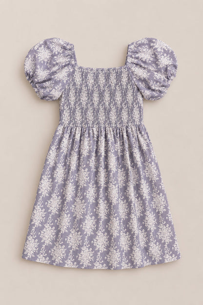Introducing the Mini Lisa Dress in Jolie Fleur by Ivy City Co: a delightful lavender dress showcasing short puff sleeves and a smocked bodice, beautifully accented with delicate white floral patterns throughout. Displayed against a neutral background, the dress exudes whimsical elegance.
