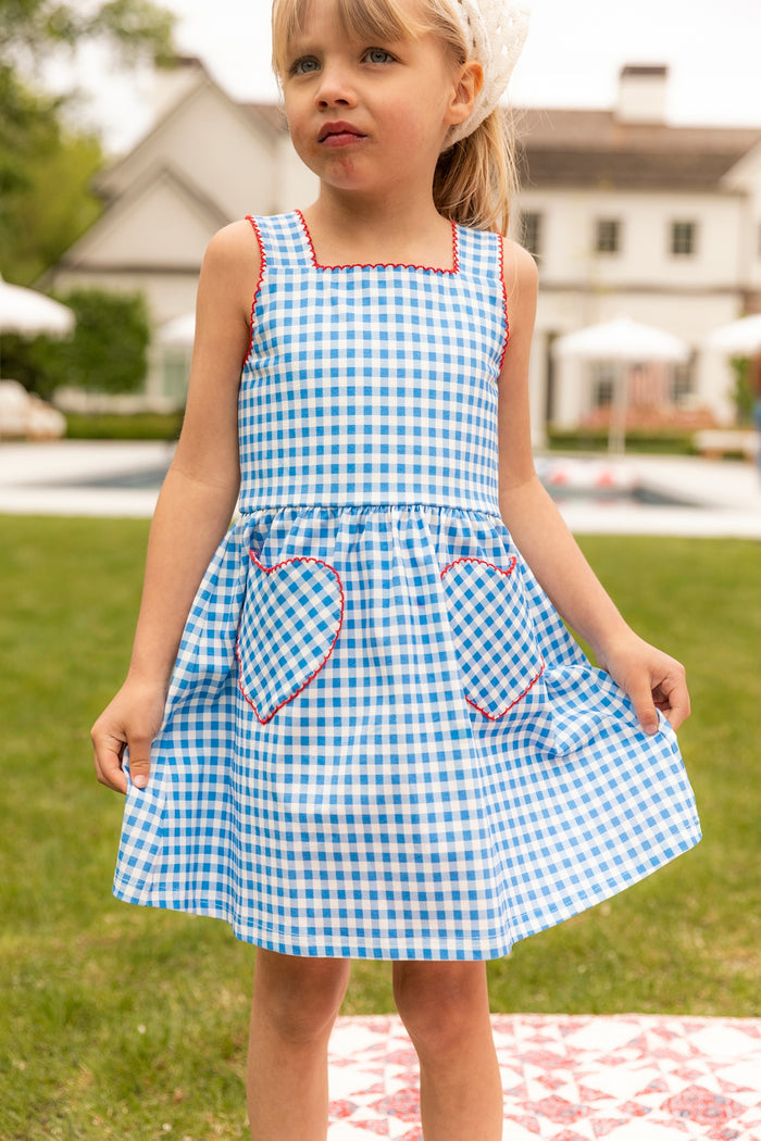 A young girl stands outdoors on grass, holding the sides of her Ivy City Co Mini Rue Play Dress in Vichy Gingham. A house and umbrella-shaded tables can be seen in the background.