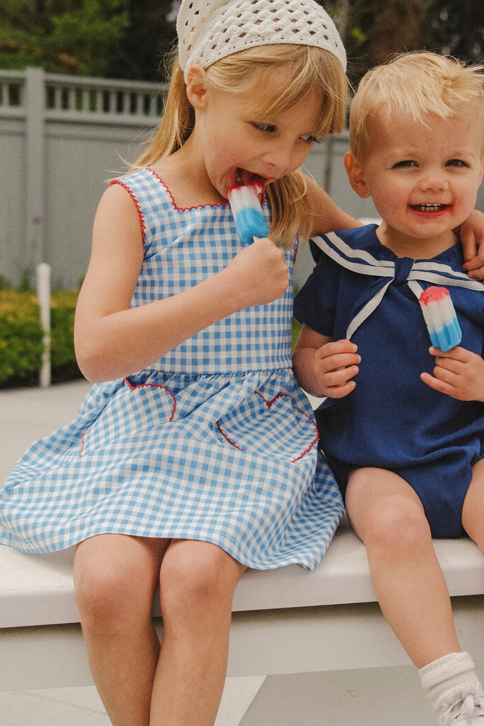 Two young children smile on a bench outdoors. The girl on the left wears the Ivy City Co Mini Rue Play Dress in Vichy Gingham and a white headscarf, enjoying a red, white, and blue popsicle beside a boy in a navy sailor outfit.