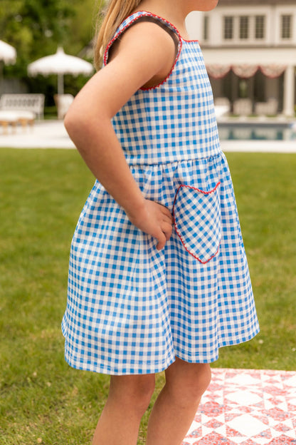 A child stands on grass outdoors, wearing the Ivy City Co Mini Rue Play Dress in Vichy Gingham, featuring blue and white checks with red trim. A house and pool are visible in the background, one hand resting on their hip.