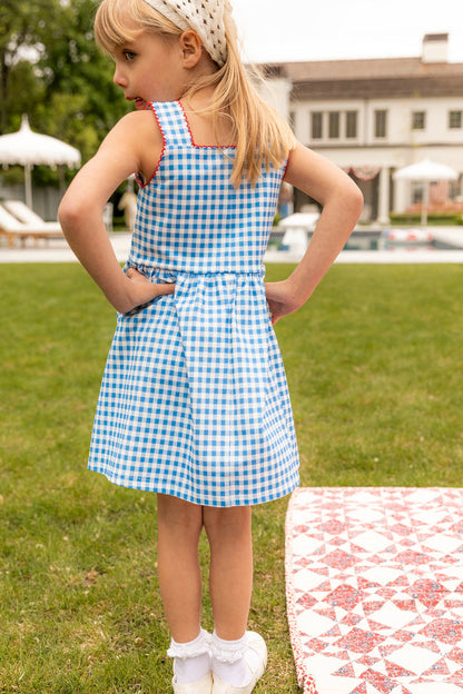 A young girl with blonde hair wears the Ivy City Co Mini Rue Play Dress in Vichy Gingham, standing on grass with hands on hips. A patterned blanket and a house with a pool are visible in the background.