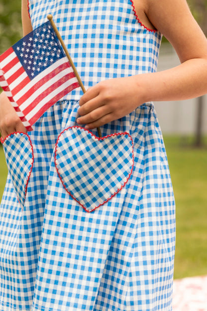A child in the Ivy City Co Mini Rue Play Dress in Vichy Gingham, featuring heart-shaped pockets, holds a small American flag outdoors on green grass with blurred trees in the background.