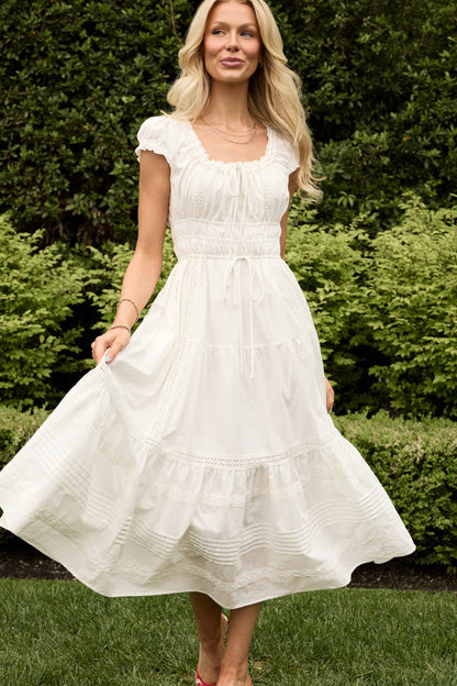 A woman in the Ivy City Co Palermo Dress in White stands on grass, holding the flowy maternity-friendly dress. She smiles, with long blonde hair and green trees and bushes behind her—perfect comfort and style for pregnancy.