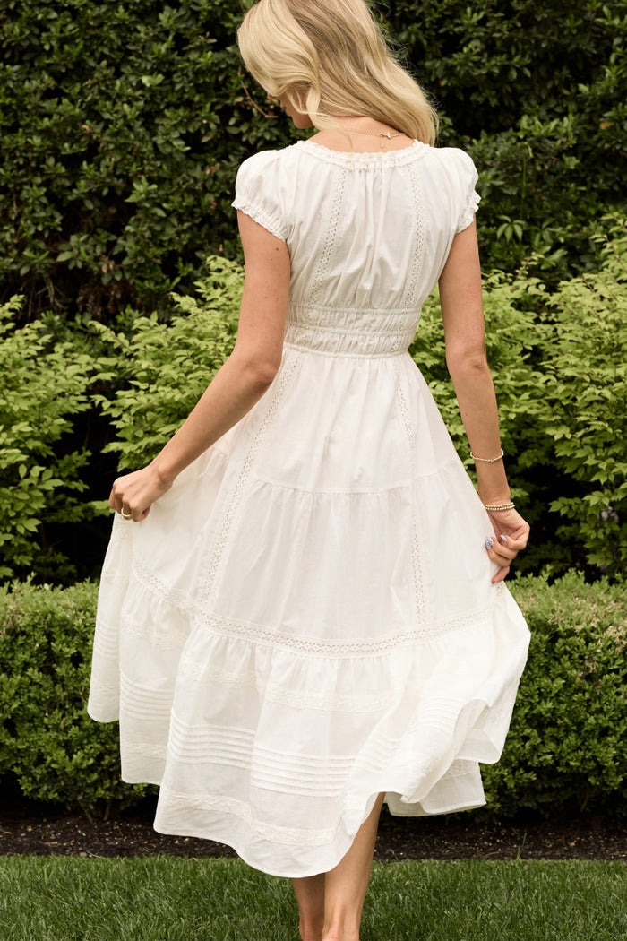 A woman wears the Ivy City Co Palermo Dress in White, standing barefoot on grass and gently twirling, surrounded by lush green bushes and trees in a garden.