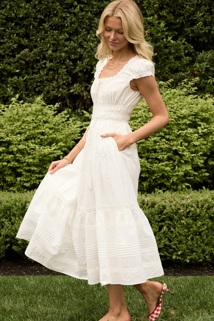 A woman with long blonde hair stands on grass, smiling and holding her skirt, wearing the Ivy City Co Palermo Dress in White. Green bushes are visible in the background.