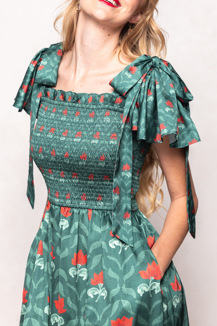 A woman wears the Ivy City Co Rayna Dress in Green Floral Block, featuring puffed sleeves tied in bows, red floral print, and a smocked bodice. This nursing-friendly style combines elegance and comfort. She has long, wavy blonde hair and red lipstick. The image is cropped to show her from shoulders to knees.