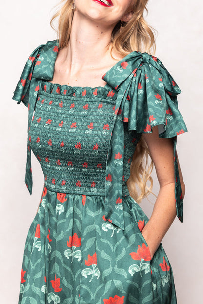 A woman wears the Ivy City Co Rayna Dress in Green Floral Block, featuring puffed sleeves tied in bows, red floral print, and a smocked bodice. This nursing-friendly style combines elegance and comfort. She has long, wavy blonde hair and red lipstick. The image is cropped to show her from shoulders to knees.