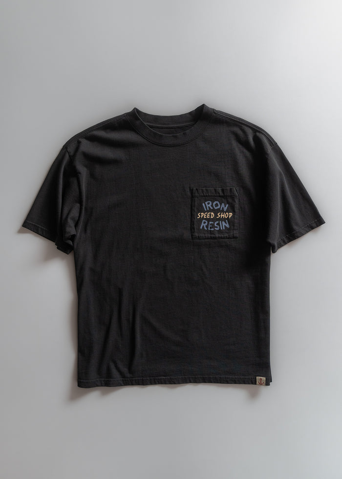 Black t-shirt with "Iron & Resin Speed Shop" on pocket, laid flat on a light gray background. Casual and simple design.