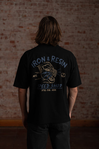 Man in black "Iron & Resin Speed Shop" shirt stands against a brick wall, showcasing the shirt's graphic design and text.