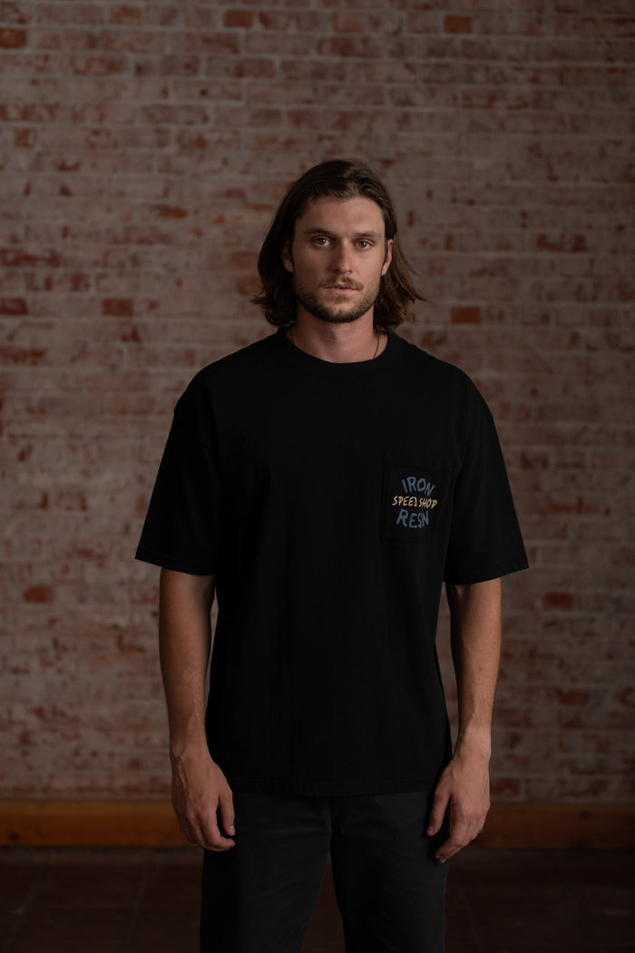 Man in a black "Iron & Resin" shirt stands against a brick wall, looking directly at the camera with a neutral expression.