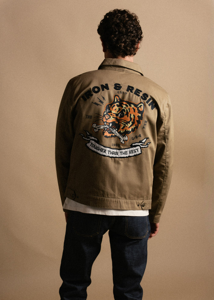 Man in brown jacket with tiger design and "Iron & Resin" text, standing against a beige background, wearing jeans.