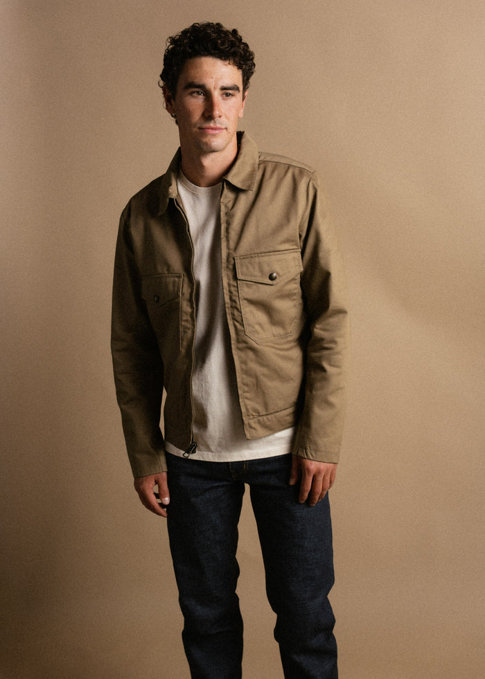 Man in a tan jacket and jeans stands against a beige background, exuding casual style and confidence.